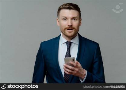Excited businessman in elegant clothes using smartphone, looking at camera with surprised face expression, male ceo executive reading good news on mobile phone while standing against grey background. Amazed businessman in suit reading good news on smartphone