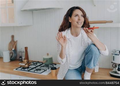 Excited caucasian young woman talking on cellphone. Happy girl with telephone sitting on worktop of table at kitchen. Modern luxurious scandinavian interior. Lifestyle concept.. Excited caucasian young woman talking on cellphone. Happy girl with telephone sitting on table.