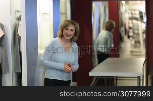 Excited charming blonde adult female choosing new dress in store fitting room. Cheerful blonde lady trying new dress in fitting room, looking in mirror while showing her new apparel to female friend. Selective focus on reflection in the mirror.