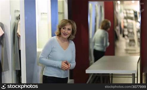 Excited charming blonde adult female choosing new dress in store fitting room. Cheerful blonde lady trying new dress in fitting room, looking in mirror while showing her new apparel to female friend. Selective focus on reflection in the mirror.
