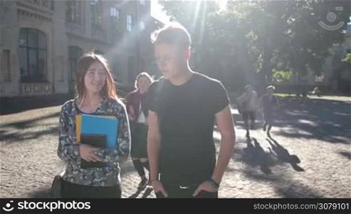 Excited college students with bags and backpacks meeting outside university building on sunny day. Joyful teenage classmates gathering on university campus while walking to college. Slow motion. Steadicam stabilized shot.