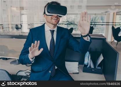 Excited entrepreneur in virtual reality glasses touching objects interact with 3D simulation, smiling male in VR goggles have fun testing new device while working in office, digital technology concept. Excited entrepreneur in virtual reality glasses touching objects interact with 3D simulation