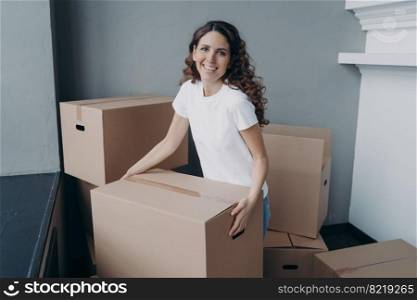 Excited female homeowner carrying boxes with things into home, settling in first new house apartment. Smiling latina girl holding carton during unpacking belongings. Happy moving day, mortgage.. Excited female homeowner holding box with things, settling in new house apartment. Happy moving day