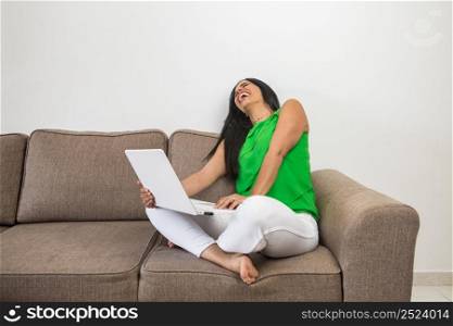 Excited female in casual clothes with netbook laughing at funny joke while sitting cross legged on couch on weekend day at home. Hispanic woman with laptop laughing at joke