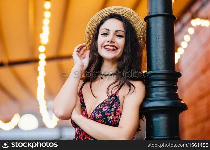 Excited female with dark hair, shinig eyes and red lips, dressed in dress and summer hat, posing at camera with happy and confident expression. Attractive female brunette with make-up resting at cafe