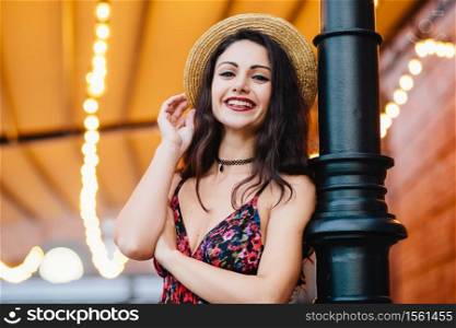 Excited female with dark hair, shinig eyes and red lips, dressed in dress and summer hat, posing at camera with happy and confident expression. Attractive female brunette with make-up resting at cafe