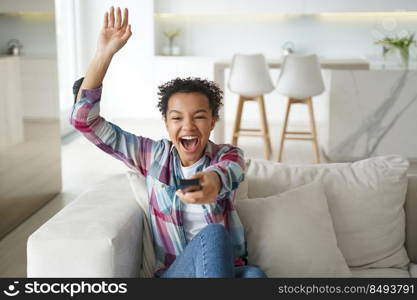 Excited football team fan is relaxing at home watching the game. African american teenage girl enjoying tv watching and shifts channels. Reality show or soccer match watching. Smart tv using.. Excited football team fan is relaxing at home watching the game. Soccer match watching.