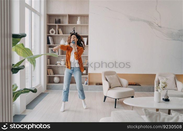 Excited girl having fun crazy in her living room. Happy european woman is singing at home with phone as with microphone. Favourite song singing. Dancing alone on weekend morning.. Excited girl having fun crazy in her living room. Happy european woman is singing at home.