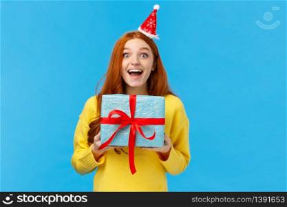 Excited girl love christmas holidays and receiving presents, holding lovely new year gift, smiling joyfully and amused, wearing santa hat as attend party, standing blue background upbeat.. Excited girl love christmas holidays and receiving presents, holding lovely new year gift, smiling joyfully and amused, wearing santa hat as attend party, standing blue background upbeat