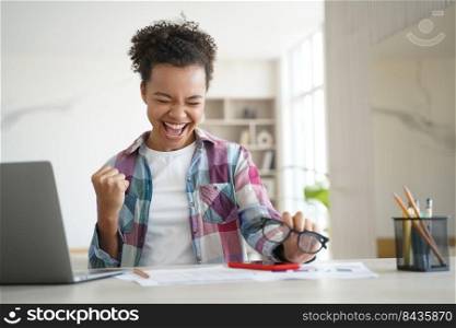 Excited happy african american high school girl, celebrating victory, got email with good exam test scores. Overjoyed biracial teen student got good news online sitting at laptop. Distance education. Excited biracial girl student celebrates victory, got email with good scores. Personal achievement