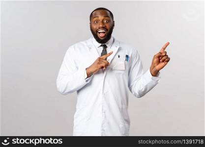Excited, happy and surprised african-american male doctor telling coworkers great news, pointing fingers upprt right corner, smiling cheerful and rejoicing, heard great news TV, grey background.. Excited, happy and surprised african-american male doctor telling coworkers great news, pointing fingers upprt right corner, smiling cheerful and rejoicing, heard great news TV, grey background