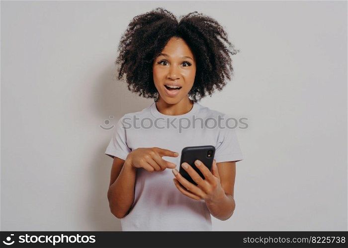 Excited happy dark skinned lady with curly hair holding mobile phone and reading good news notification, enjoying chatting with friends or doing online shopping, isolated on white studio background. Excited happy dark skinned lady with curly hair holding mobile phone