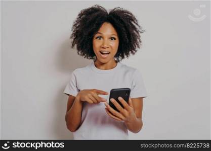 Excited happy dark skinned lady with curly hair holding mobile phone and reading good news notification, enjoying chatting with friends or doing online shopping, isolated on white studio background. Excited happy dark skinned lady with curly hair holding mobile phone