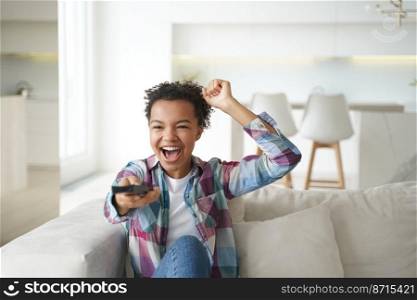 Excited happy mixed race young girl watching football on tv, screaming, makes yes winner gesture. Energetic young teen lady sport fan celebrates favorite team victory, sitting on couch at home.. Excited mixed race young girl sport fan watching football on tv, screaming, makes yes winner gesture