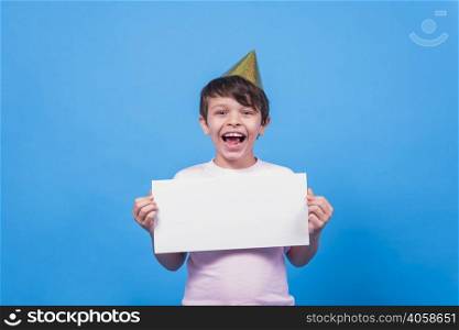 excited little boy wearing party hat holding blank card hand blue surface