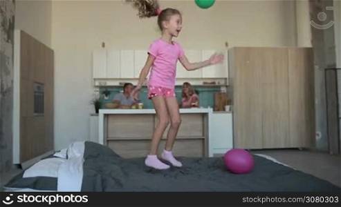 Excited little girl in pajamas jumping with baloons on bed and smiling while parents having breakfast in kitchen on background. Lovely child enjoying great time at home, laughing and having fun over domestic interior background. Slo mo. Dolly shot.