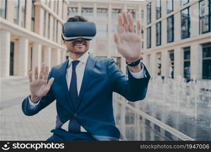 Excited male office worker in formal suit checking information in virtual reality using modern VR glasses while sitting outside on bench, happy businessman interacting with cyberspace. Excited office worker in formal suit checking information in virtual reality