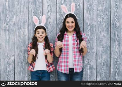 excited mother daughter with bunny ears pointing finger downward against wooden backdrop