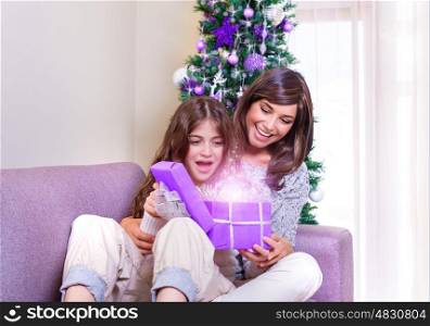 Excited mother with daughter sitting at home near beautiful decorated Christmas tree, enjoying magical gift box, New Year fairy tale concept