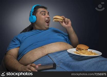 Excited obese man with belly visible out of his shirt eating burger while listening to music with a plate of burger and french fries on his thigh