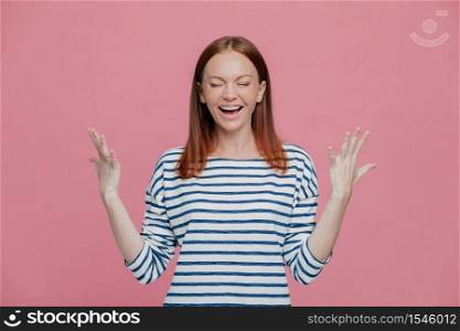 Excited overjoyed woman closes eyes, keeps hands raised, dressed in striped sweater, laughs positively, isolated over pink background. People, happiness, joy concept. Glad young female model