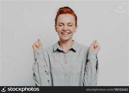 Excited overjoyed young ginger female raising hands with clenched fists and keeping eyes closed, making yes gesture while posing isolated on grey studio background. Success concept. Excited overjoyed young ginger female raising hands with clenched fists, keeping eyes closed