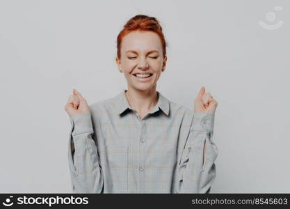 Excited overjoyed young ginger female raising hands with clenched fists and keeping eyes closed, making yes gesture while posing isolated on grey studio background. Success concept. Excited overjoyed young ginger female raising hands with clenched fists, keeping eyes closed