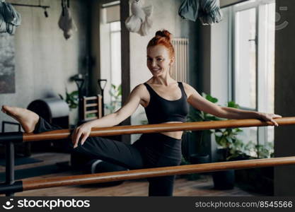 Excited slim red haired woman standing in modern fitness studio with her hands and one leg on ballet barre, doing stretching exercises and looking into camera, wearing comfortable black sportswear. Happy young woman good stretch posing in fitness studio during barre workout