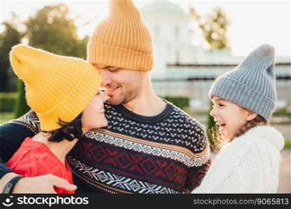 Excited small kid looks at her parents who are in love, going to kiss each other, have goos relationships, embrace. Affectionate woman and man support. Smiling little girl looks at father and mother