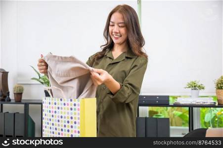 Excited smile female shopper holding ordered shirt, Happy Asian young customer woman opening shopping bag package after ordered new fashion clothes at home, shopping online delivery shipping