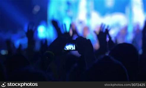 Excited viewers applauding to the singer or band performing on colored illuminated stage. One of the fans taking picture or making video with digital camera