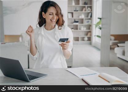 Excited woman holding bank credit card uses online banking service at laptop. Happy female makes yes gesture, pleased with cashback, easy successful payment. E-banking, e commerce.. Female holds bank credit card with cashback using online banking service at laptop makes yes gesture