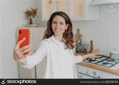 Excited woman holding smartphone, making video call in modern kitchen at home, looking at phone screen, happy female food blogger, showing oven, chatting online with followers in social networks.. Female food blogger holding phone, shows kitchen, chatting online with followers, making video call