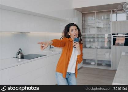 Excited woman in earphones listening to music, singing using smartphone as mic, dancing in kitchen at home. Energetic hispanic girl sings pointing by finger ahead. Musical mobile apps advertising.. Energetic girl listens to music, singing using smartphone as mic at home. Musical mobile apps ad