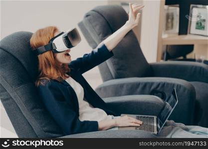 Excited woman in VR headset and laptop, touching air during virtual reality experience while sitting in chair in internet cafe salon. Emotional lady exploring artificial 3D world or playing video game. Excited woman in VR headset and laptop, touching air with hand, sitting in armchair in coffee shop