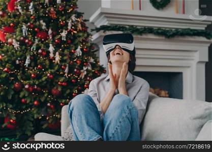 Excited woman in VR headset glasses enjoying Christmas holidays at home, laughing female wearing virtual reality goggles relaxing on cozy sofa in living room decorated for Xmas, playing 3D games. Overjoyed woman in VR headset sitting on sofa in living room with Xmas tree on background