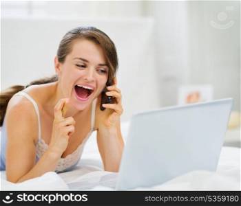 Excited woman with laptop speaking mobile with crossed fingers