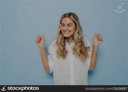 Excited young blonde girl filled with joy cheering with both hands in air, can not contain her happiness expressing her emotions with gestures while posing isolated over blue wall. Excitement concept. Blonde girl with closed eyes cheering with both hands in air