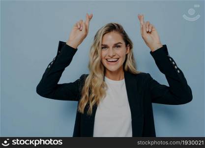 Excited young blonde woman in black coat waiting for special moment, keeping fingers crossed in anticipation of wish to come true, standing isolated in front of blue background in studio. Excited blonde girl wishing with crossed fingers