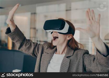 Excited young business woman experiencing virtual reality using VR glasses while standing in office, expressing emotions with gesture and happy facial expression, cannot believe what she is seeing. Excited young business woman experiencing virtual reality using VR glasses at work