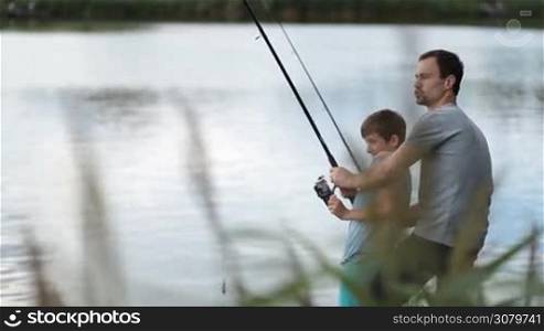 Excited young dad and teenage boy pulling fish out from freshwater pond together over blue water surface background. Joyful father helping son to fight a fish, they caught on bait at the lake while family angling with fishing rod and reel. Side view.