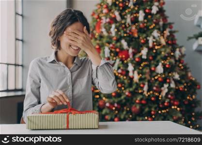 Excited young european woman getting christmas surprise, sitting at desk near decorated xmas tree at home, happy hispanic female covering her eyes by hand and opening wrapped New Year gift box. Excited european woman getting christmas surprise, sitting at desk near decorated xmas tree at home