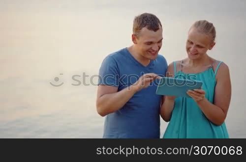 Excited young man and woman by the sea discussing something shown on touch pad
