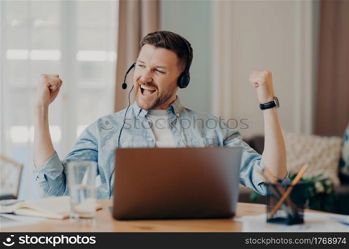 Excited young man freelancer headset with microphone celebrating sucess after important online meeting or job interview, sitting at desk in living room at home. Winning behaviour concept. Caucasian bearded guy enjoying successful negotiations online