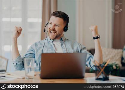 Excited young man freelancer headset with microphone celebrating sucess after important online meeting or job interview, sitting at desk in living room at home. Winning behaviour concept. Caucasian bearded guy enjoying successful negotiations online