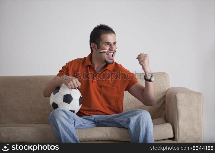 Excited young man with soccer ball cheering while sitting on sofa at home