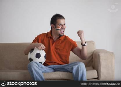 Excited young man with soccer ball cheering while sitting on sofa at home