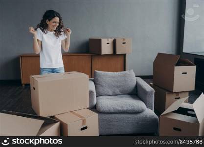 Excited young woman is packing things. Spanish girl moves to new house. Lady is unpacking cardboard boxes in new apartment. Big new living room with furniture. Real estate purchase concept.. Excited young woman is packing things. Spanish girl moves to new house. Real estate purchase.