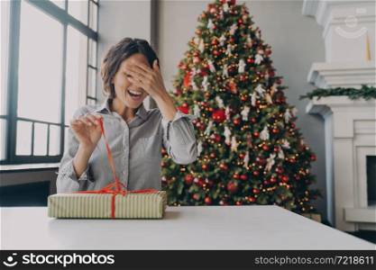Excited young woman sitting at table in living room decorated with beautiful bright Xmas tree, joyful lady unpacking Christmas gift with one hand and with another closing her eyes to being surprised. Excited young woman unpacking Christmas gift while sitting in room decorated with bright Xmas tree