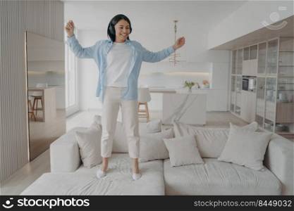 Excited young woman wearing wireless headphones jumping on sofa alone at home. Joyful energetic female having fun, enjoy musical sound, dancing listening to music on couch in living room on weekend.. Excited energetic woman wearing headphones having fun enjoying music jumping on sofa alone at home